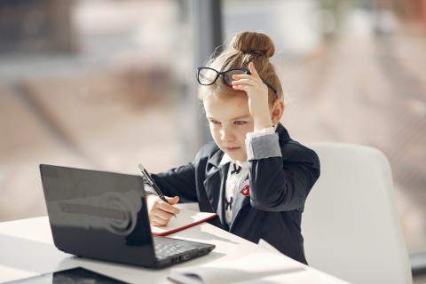 Bildrecht: istock Ein kleines Mädchen in Businesskleidung sitzt konzentriert an einem Schreibtisch mit Laptop und Notizbuch. Sie hebt ihre Brille an die Stirn, hält einen Stift in der Hand und wirkt fokussiert, als ob sie arbeitet oder etwas Wichtiges plant.