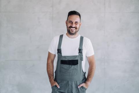 Bildrecht: istock Ein lächelnder Mann in grauer Latzhose und weißem T-Shirt steht vor einer hellgrauen Betonwand und hat die Hände in den Hosentaschen.